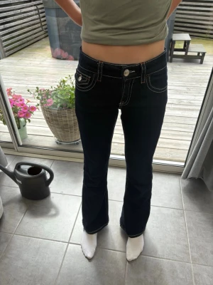Mörkblå bootcut jeans med kontrastsömmar - Säljer ett par mörkblå bootcut jeans med vita kontrastsömmar och dekorativa fickor både fram och bak. Jeansen har låg midja och stängs med knapp och dragkedja. Perfekta för dig som gillar en klassisk men ändå trendig look.