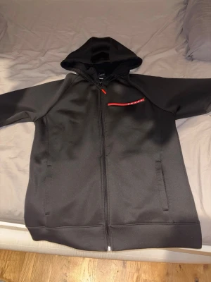 Svart hoodie från Prada med dragkedja - Snygg svart hoodie från Prada med hel dragkedja framtill och huva. Diskret röd detalj med Prada-logga på bröstet. Tillverkad i ett mjukt och lite glansigt material som känns lyxigt. Perfekt för en stilren och modern look.