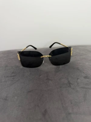 Svarta solglasögon från Miu Miu - Snygga solglasögon från Miu Miu med svarta, rektangulära glas och tunna guldfärgade metallbågar. På sidan av glasen sitter Miu Miu-loggan i guld. Glasögonen har en modern och edgy look som sticker ut.