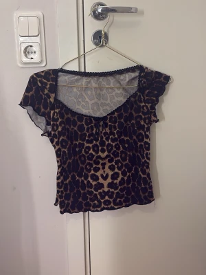 Leopardmönstrad topp med rosett - Snygg leopardmönstrad topp med korta volangärmar och liten rosett framtill. Toppen har en lätt transparent känsla och dekorativ spetskant vid ringningen. Perfekt för dig som vill sticka ut med ett djurmönster.