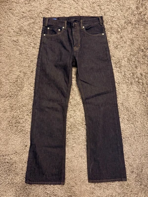 Mörkblå raka jeans i rådenim - Säljer ett par mörkblå jeans i rådenim med rak passform och klassisk femficksdesign. Jeansen har kontrastsömmar och silverfärgade nitar. Perfekta för dig som gillar en clean och tidlös look. Materialet är kraftig denim i bomull.