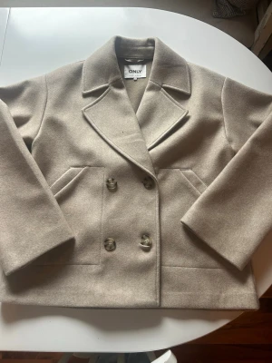 Beige kappa från ONLY med bred krage - Säljer en stilren beige kappa från ONLY med dubbelknäppning och breda slag. Kappan har två framfickor och stora knappar i matchande färg. Nypris 999 sek. Använd enstaka gånger.