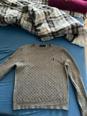 Grå kabelstickad tröja Polo Ralph Lauren - Snygg grå kabelstickad tröja från Polo Ralph Lauren med rund halsringning och klassisk broderad logga på bröstet. Tröjan har långa ärmar och ribbade muddar vid ärmslut och nederkant. Perfekt för dig som gillar stilren och tidlös design.