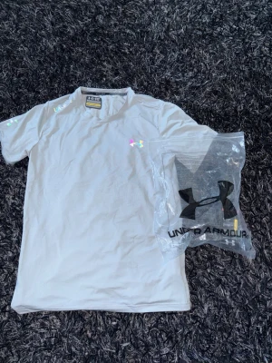 Grå t-shirt från Under Armour - Snygg vit t-shirt från Under Armour med klassisk logga på bröstet. T-shirten har korta ärmar och är tillverkad i ett mjukt och funktionellt material som passar perfekt till träning. Enkel och stilren design med diskreta detaljer.