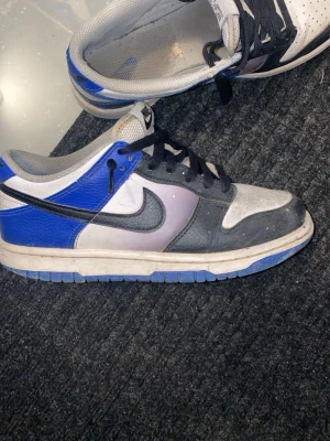 Nike Dunk Low blå/svart/vit - Säljer ett par Nike Dunk Low sneakers i blått, svart och vitt med klassisk Swoosh på sidan. Skorna har lågt skaft, svart snörning och en slitstark yttersula. Perfekta för dig som gillar streetwear och vill ha en ikonisk sneaker i din samling.