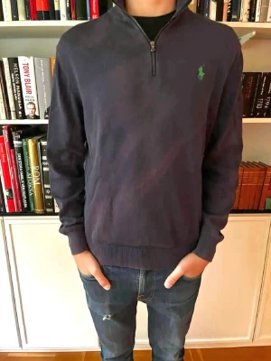 Mörkblå halvzip från Ralph Lauren - Mörkblå tröja med halv dragkedja och hög krage från Ralph Lauren. Klassisk grön logga broderad på bröstet och ribbade muddar vid ärmslut och nederkant. Perfekt till jeans för en clean och stilren look.