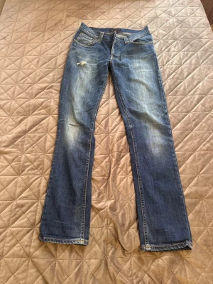 Blå bootcut jeans från Nudie Jeans - Säljer ett par klassiska blå bootcut jeans från Nudie Jeans med slitningar och en liten reva på vänster lår. Jeansen har fem fickor, orangea sömmar och knappgylf. Perfekta för en avslappnad och cool stil.