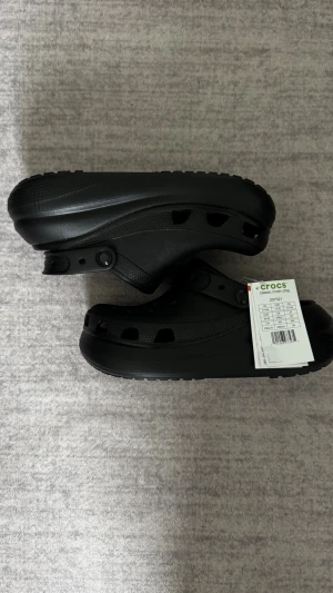 Crocs Classic Crush Clog - Svarta Crocs Classic Clog med ventilerande hål och justerbar hälrem. Tillverkade i lätt och bekvämt syntetmaterial som är enkelt att rengöra. Storlek M7 eller W9 vilket motsvarar EU40.