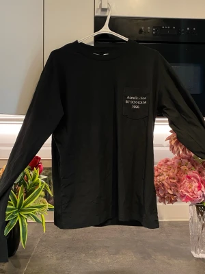 Svart långärmad topp Acne Studios - Säljer en svart långärmad topp från Acne Studios med bröstficka och vit broderad text 'Acne Studios STOCKHOLM 1996' på fickan. Klassisk passform och rund halsringning, perfekt till alla dina outfits.