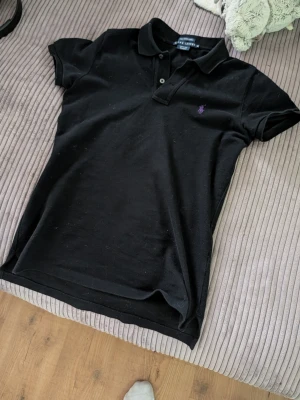 Svart pikétopp från Ralph Lauren - Klassisk svart pikétopp från Ralph Lauren med lila broderad logga på bröstet. Modellen har korta ärmar, krage och knappar framtill. Perfekt till jeans eller kjol för en clean och sportig look.