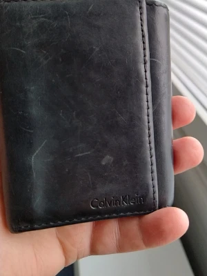 Svart läderplånbok Calvin Klein - Stilren svart plånbok från Calvin Klein i äkta läder. Plånboken har en klassisk, fyrkantig form och diskret logga präglad på framsidan. Perfekt för dig som gillar minimalistisk design och vill ha en smidig plånbok i fickan.