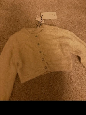 Fluffig cropped kofta - Supersöt beige cropped kofta med fluffig textur och rund hals. Koftan har långa ärmar och knäpps med små silvriga knappar framtill. helt ny, snabb frakt 