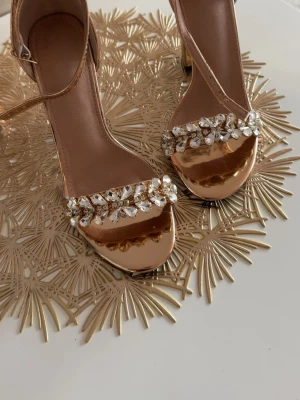 Guldiga pumps med strassdetaljer - Säljer ett par glansiga guldiga pumps med bred klack och öppen tå. Skorna har en rem runt ankeln och är dekorerade med stora, gnistrande strass på tåbandet. Perfekta för dig som vill sticka ut och addera lite extra bling till din outfit.