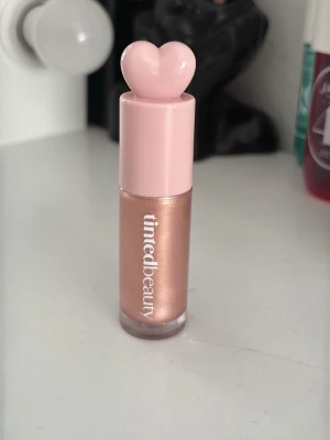 Tinted Beauty läppstift med hjärtlock - Supersött läppstift från Tinted Beauty i en skimrande roséguldig nyans. Förpackningen har ett rosa lock med ett hjärta på toppen, vilket gör den extra gullig. Perfekt för dig som gillar en glansig finish och vill ha något som sticker ut i sminkväskan.advänd 3 gångar 