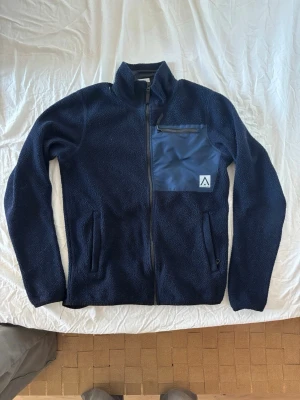 Marinblå Arket fleecejacka med dragkedja - Mysig marinblå fleecejacka från Arket med hel dragkedja framtill. Jackan har en bröstficka i kontrasterande material med dragkedja och två sidofickor. Ståkrage och diskret logga på bröstet. Perfekt för lager-på-lager och kyliga dagar.