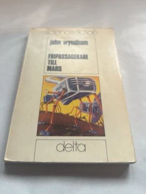 Fripassagerare till Mars - Wyndham - Science fiction-bok med titeln 'Fripassagerare till Mars' av John Wyndham. Omslaget är beige med en färgstark illustration av en futuristisk maskin på Mars och tydlig titeltext. Pocketutgåva från förlaget Delta.