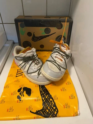 Nike x Off-White Dunk Low sneakers - Unika Nike x Off-White Dunk Low sneakers i vitt och grått med olivgröna och vita snören, orange detaljer och Off-White zip-tie. Skorna har perforerad tåbox, dubbla snörningar och klassisk Off-White design. Kommer med originalkartong och specialförpackning. Perfekt för dig som vill sticka ut med klass. Låda tillkommer såklart. För mer frågor är det bara att höra av sig i dm. 