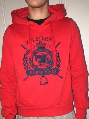 Röd Falsterbo Horse Show hoodie - Snygg röd hoodie från Falsterbo Horse Show med stort blått tryck framtill. Klassisk huva med dragsko och ribbade muddar. Perfekt för dig som gillar hästsport och vill ha en sportig och bekväm stil. Tillverkad i mjukt material. I storlek M men funkar som S funkar oxå för män och kvinnor.