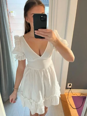 Vit kort omlottklänning med puffärm - Supersöt vit kort klänning med puffärmar och omlott-v-ringning. Kjolen har volangkant och klänningen sitter åt i midjan för en feminin siluett. Perfekt för sommarens alla tillfällen och ger en romantisk vibe.
