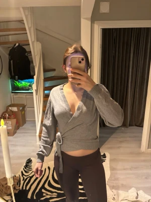 Grå omlottstickad tröja - Säljer en grå stickad omlottopp med långa ärmar och knytband i midjan. Tröjan har ribbade muddar och en djup v-ringning som ger en trendig look. Perfekt att styla med både jeans och kjol.