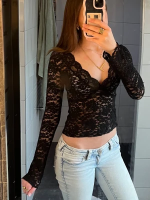 Svart spetsblus med v-ringning - Säljer en svart, långärmad blus i spets med v-ringad hals och croppad passform. Blusen har fina spetsdetaljer och transparenta partier som ger en trendig look. Perfekt att styla med jeans eller kjol för en cool outfit.