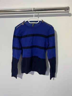 Blå och svart stickad tröja Ralph lauren - Snygg blå stickad tröja från Ralph Lauren med svarta horisontella ränder och detaljerade dragkedjor på axlarna. Tröjan har lång ärm och rund halsringning. Perfekt för dig som gillar en stilren men ändå unik look.