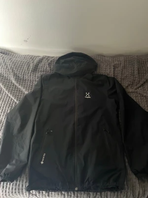 Svart skaljacka från Haglöfs - Snygg svart skaljacka från Haglöfs med huva och dragkedja framtill. Jackan har två fickor med dragkedja och är tillverkad i slitstarkt GORE-TEX-material. Perfekt för blåsiga dagar och outdoor-äventyr. Diskret Haglöfs-logga på bröstet.