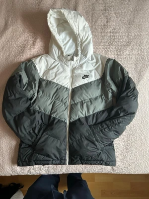 Nike pufferjacka i vitt och grått - Säljer en snygg pufferjacka från Nike i vitt och olika nyanser av grått. Jackan har huva, dragkedja framtill och den klassiska Nike-loggan på bröstet. Perfekt för kalla dagar och riktigt skön att bära. Materialet är syntet och jackan har en sportig look.