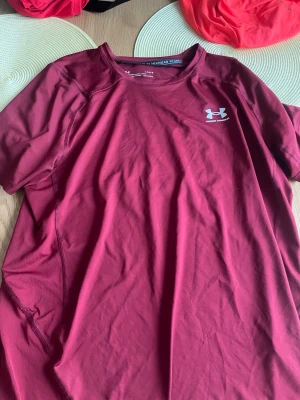 Vinröd träningströja Under Armour - Snygg vinröd träningströja från Under Armour med korta ärmar och rund hals. Tillverkad i ett lätt och snabbtorkande funktionsmaterial, perfekt för träning och sportaktiviteter. Diskret logga på bröstet.