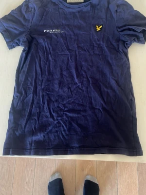 Mörkblå t-shirt från Lyle & Scott - Snygg mörkblå t-shirt från Lyle & Scott med klassisk gul logga på bröstet och tryckt text. Tillverkad i mjuk bomull och har en normal passform med korta ärmar. Perfekt till jeans eller shorts för en clean look.