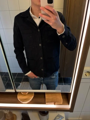 Svart overshirt från Lexington - Stilren svart overshirt från Lexington i kraftig bomull. Klassisk rak passform med fyra fickor framtill och knäppning med metallknappar. Perfekt att bära över en t-shirt eller skjorta för en avslappnad men snygg look.