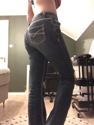 Mörkblå slitna bootcut jeans - Säljer ett par mörkblå bootcut jeans med slitningar och hål vid knäna. Jeansen har kontrastsömmar och bakfickor med dekorativa sömmar. Perfekta för en avslappnad och trendig look.