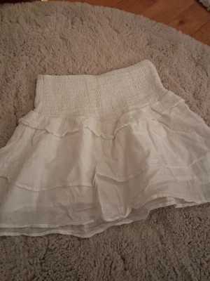 Vit volangkjol från Gina Tricot - Supersöt vit kjol från Gina Tricot med bred smock i midjan och dubbla volanglager. Kjolen är gjord i ett lätt och luftigt tyg som ger en flowy känsla. Perfekt för dig som gillar en romantisk och trendig stil.