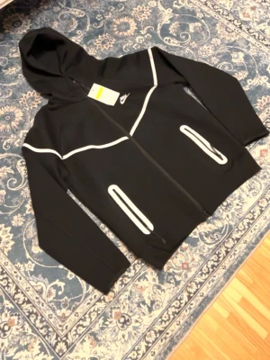 Svart Nike hoodie med reflexdetaljer - Snygg svart hoodie från Nike med dragkedja och vita reflexdetaljer på bröstet och vid fickorna. Hoodien har två fickor med dragkedja och en klassisk huva. Perfekt för dig som vill sticka ut och synas i mörkret. Tillverkad i mjukt och bekvämt material.