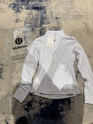 Vit träningsjacka från Lululemon - Snygg vit träningsjacka från Lululemon med figurnära passform, hel dragkedja framtill och två fickor med dragkedja. Tillverkad i ett stretchigt funktionsmaterial som andas. Perfekt för löpning eller gym.