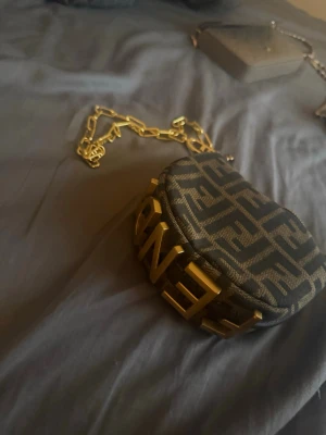 brun monogram handväska - Säljer en brun handväska. Väskan har en chunky guldfärgad kedja och stora guldbokstäver framtill. Perfekt för dig som gillar statement-accessoarer och exklusiv design.