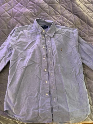 Blå skjorta från Ralph Lauren - Klassisk ljusblå skjorta från Ralph Lauren med button-down krage och broderad logga på bröstet. Skjortan har långa ärmar och vita knappar framtill. Perfekt till jeans eller chinos för en clean och stilren look.