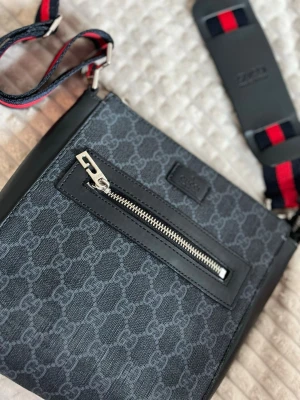 Svart axelväska från G - Snygg svart axelväska från G med klassiskt GG-mönster i grått. Väskan har en framficka med dragkedja och detaljer i svart läder. Axelremmen är justerbar och har röd-blå ränder för en sportig touch. Perfekt för dig som vill ha en stilren och trendig väska.