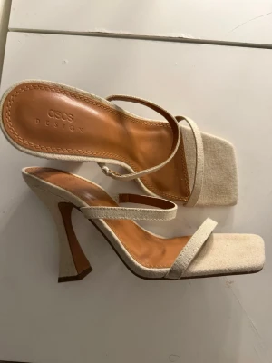 Beige pumps med fyrkantig tå ASOS - Stilrena pumps från ASOS DESIGN i beige tyg med två smala remmar och fyrkantig tå. Klackarna är höga och har en lätt utsvängd form. Innersulan är i brunt konstläder för extra komfort. Perfekta för dig som gillar trendiga och minimalistiska skor.