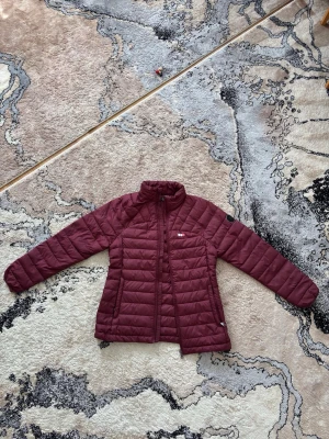 Vinröd pufferjacka från True North  - Snygg vinröd pufferjacka från True North med hög krage och dragkedja framtill. Jackan har två sidofickor med dragkedja. Perfekt för kalla dagar och ger en clean look. Materialet är lätt och isolerande syntet. Anledning till att jag säljer är på grund av att jag har flera andra jackor som används istället. 