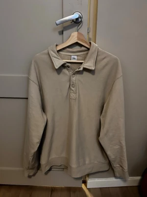 Beige collagetröja från Zara - Snygg beige collagetröja från Zara med klassisk krage och knappar vid halsen. Tillverkad i mjukt sweatshirtmaterial som känns skönt mot huden. Perfekt för en avslappnad och stilren look.