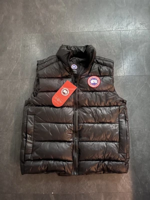 Canada Goose väst - Snygg svart dunväst från Canada Goose med hög krage och klassisk logotyp på bröstet. Västen har en quiltad design, dragkedja framtill och två sidofickor. Perfekt för lager-på-lager och håller dig varm under kyliga dagar.