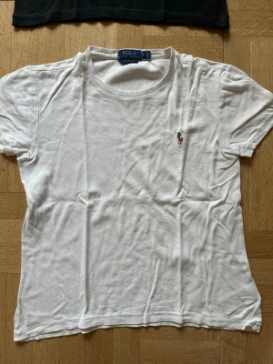 Vit t-shirt från Polo Ralph Lauren - Klassisk vit t-shirt från Polo Ralph Lauren med korta ärmar och rund hals. Liten broderad logga på bröstet i färg. Tillverkad i mjuk bomull för en skön och avslappnad look. Perfekt basplagg till garderoben.