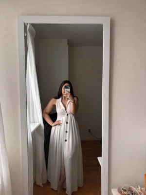 Vit ärmlös långklänning med knappar - Säljer en stilren vit långklänning med djup v-ringning och stora knappar framtill. Klänningen är ärmlös, har markerad midja och lätt utställd kjol med slits. Perfekt för sommarens alla tillfällen.