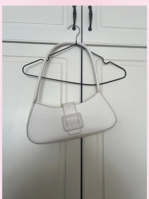 Vit axelväska med spänne - 💓Snygg vit axelväska i minimalistisk design med ett stort dekorativt spänne framtill. Väskan har en mjukt böjd form och långa handtag som gör den enkel att bära över axeln. Materialet ser ut som konstläder och väskan har en stilren look som passar till många outfits.💞fler väskor ser du i min sida