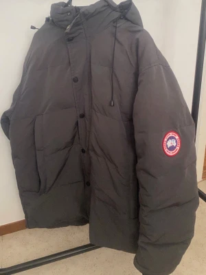 Grå dunjacka från Canada Goose - Säljer en grå dunjacka från Canada Goose med huva och dragsko. Jackan har stora fickor framtill och den klassiska Canada Goose-loggan på ärmen. Perfekt för kalla vinterdagar och riktigt varm tack vare dunfyllningen.