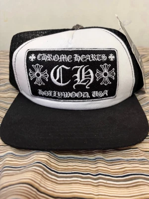 Chrome Hearts truckerkeps  - Säljer en svart och vit truckerkeps från Chrome Hearts med broderad logga och texten 'Hollywood, USA' framtill. Kepsen har mesh på sidorna och justerbar snapback. Märkestaggar sitter kvar. Perfekt för dig som gillar streetwear och vill sticka ut.