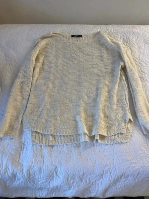 Krämvit stickad tröja - Mysig krämvit stickad tröja med rund halsringning och långa ärmar. Tröjan har en avslappnad passform, ribbstickade muddar och en liten slits i sidorna. Perfekt för lager på lager och enkel att matcha med jeans eller kjol.