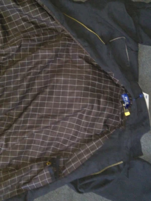 Svart vindjacka med rutigt foder - Snygg svart vindjacka med gulddragkedja och rutigt innerfoder i brunt och vitt. Jackan har en klassisk krage och elastisk nederkant. Perfekt för blåsiga dagar och enkel att matcha med olika outfits. Liten L