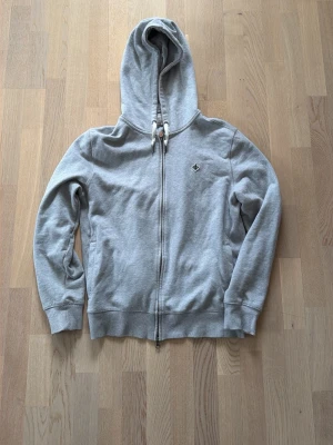 Grå zip hoodie Morris - Snygg grå hoodie från Morris med dragkedja framtill och broderad logga på bröstet. Tröjan har huva med vita dragsnören och ribbade muddar vid ärmar och nederkant. Perfekt för en chill och avslappnad stil.